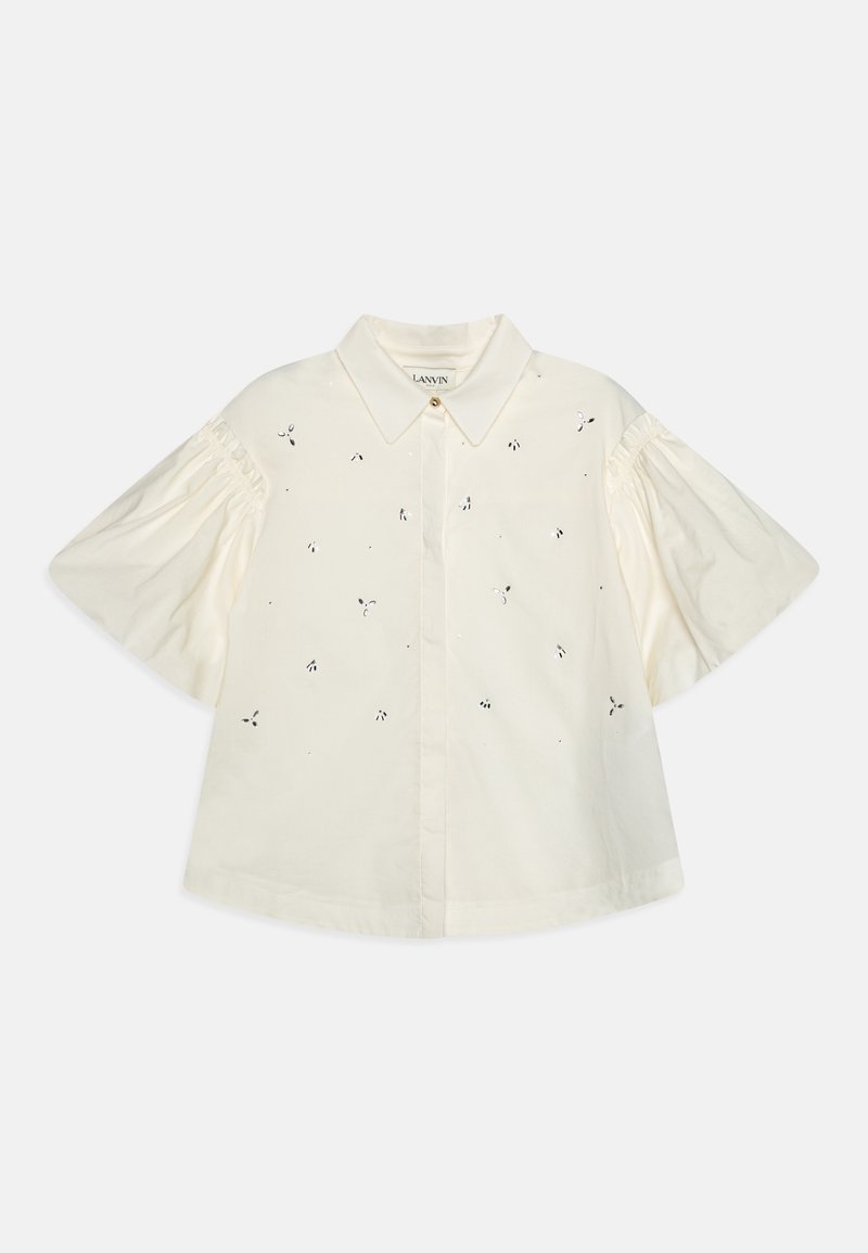 Blouse blanche à manches courtes avec col pointu, manches bouffantes et petits motifs découpés éparpillés.