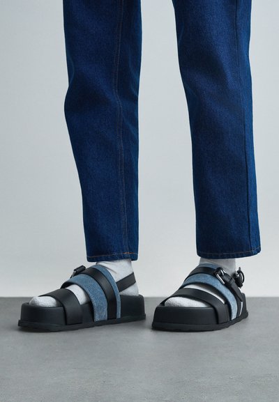 Férfi fehér zokniban, fekete és farmerszíjjal készült szandálban és kék farmerben áll egy szürke padlón, egy egyszínű háttér előtt.