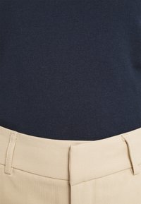 Lähedalt vaade beezile kõrge vöökohtadega pükste vööle, millele on sätitud tumesinine kootud topp.