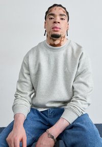 Sudadera de algodón gris con puños de canalé y cuello redondo, con un pequeño logo blanco. Combinada con jeans de mezclilla azul.