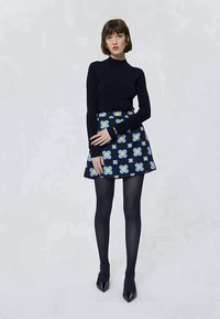 Frau in schwarzem Rollkragenpullover, marineblauer Rock mit blauem und gelbem Blumenmuster, schwarzen Strumpfhosen und schwarzen spitzen Schuhen, stehend vor einfarbigem Hintergrund.
