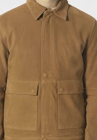 Brązakowa kurtka z dwiema kieszeniami, wykonana z teksturowanej tkaniny, z zapięciem na guziki i spiczastym kołnierzem. Projekt ma prosty krój i gładkie wykończenie.