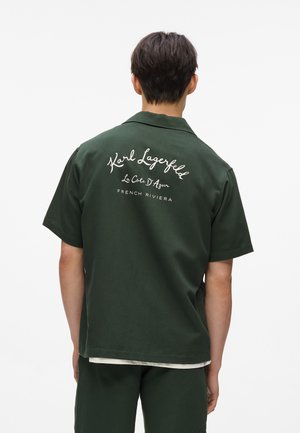 Grünes kurzärmliges Hemd aus Stoff mit aufgesticktem weißem Text auf der Rückseite, der "Karl Lagerfeld La Côte D'Azur FRENCH RIVIERA" lautet.