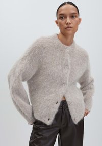 Woman wearing a fuzzy light gray buttoned cardigan paired with black high-waisted leather pants, posed against a plain light background.