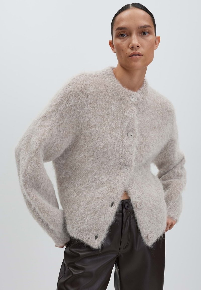 Woman wearing a fuzzy light gray buttoned cardigan paired with black high-waisted leather pants, posed against a plain light background.