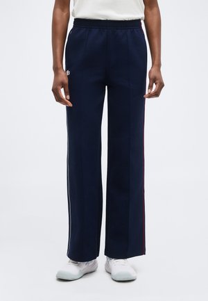 Personne portant un pantalon droit bleu marine avec des bandes latérales, des baskets blanches et une chemise blanche, debout devant un fond uni.