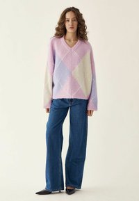 V-Ausschnitt, übergroßer Pullover im pastellfarbenen Pink-, Lila- und weißen Karomuster. Weicher, strukturierter Stoff. Kombiniert mit hochtaillierten blauen Jeans.