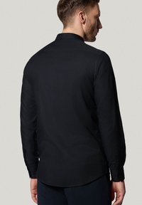Czarna koszula z długim rękawem, z wywijanym kołnierzykiem, gładką fakturą, zapinanymi mankietami i zaokrąglonym dołem, widziana z tyłu.