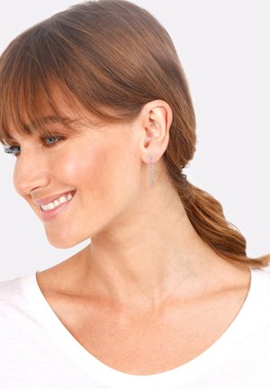 Boucles d'oreilles en argent avec un design de chaîne fine et pendante. Des pierres claires près du lobe d'oreille ajoutent de l'éclat. Texture lisse et style minimaliste.