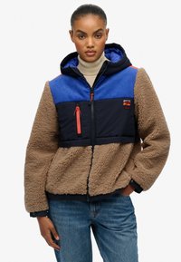 Fleecejack met een marineblauw en blauw paneelontwerp, bruine fluffy textuur, rits aan de voorkant en een capuchon. Bevat oranje accenten en een borstzak.