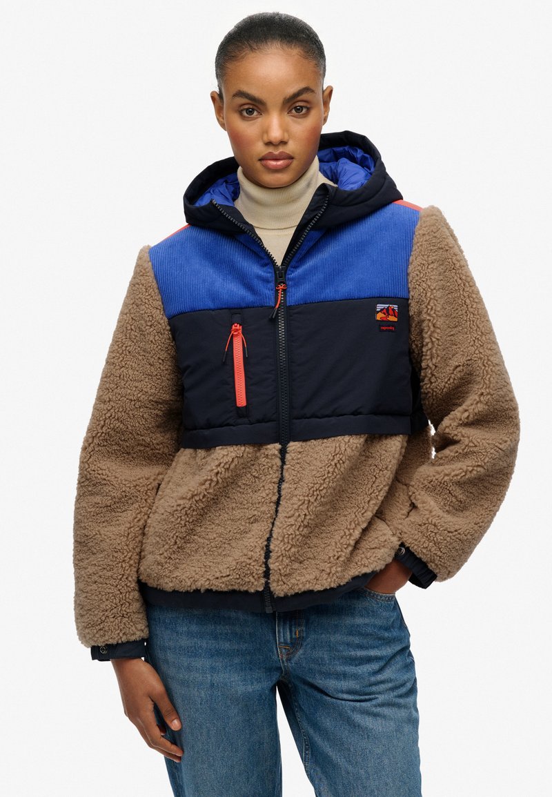 Fleecejack met een marineblauw en blauw paneelontwerp, bruine fluffy textuur, rits aan de voorkant en een capuchon. Bevat oranje accenten en een borstzak.