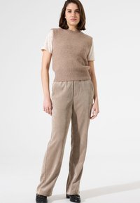 Beige gestrickter ärmelloser Pullover über einem weißen kurzärmligen Shirt, kombiniert mit hellbraunen Cordhosen. Das Modell steht vor einem weißen Hintergrund.