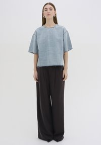 Top in denim bluastro con una forma squadrata e bordi sfrangiati, abbinato a pantaloni marroni larghi a vita alta, su uno sfondo neutro.