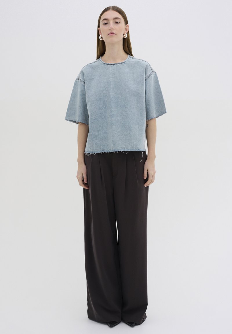 Top in denim bluastro con una forma squadrata e bordi sfrangiati, abbinato a pantaloni marroni larghi a vita alta, su uno sfondo neutro.