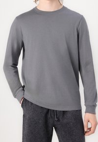 Person trägt ein schlichtes graues Langarmshirt mit Rundhalsausschnitt und schwarze Jogginghosen mit Kordelzug, eine Hand in der Tasche, vor einem schlichten Hintergrund.