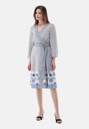 Frau trägt ein knielanges Wickelkleid mit blauen Blumen- und geometrischen Mustern, langen Ärmeln, gebunden in der Taille, und schwarzen offenen Sandalen.