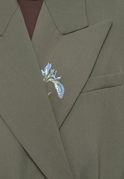 Blazer verde oliva con colletto a rever e un ricamo floreale decorativo blu e verde sulla patta. Tessuto texturizzato con vestibilità strutturata.