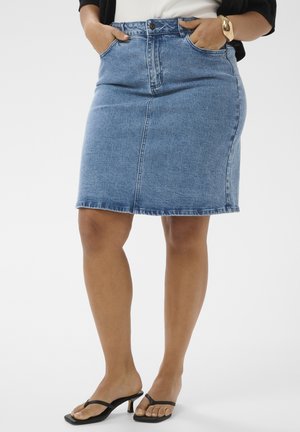 Frau trägt einen knieumspielenden blauen Jeansrock, ein schwarzes kurzärmeliges Oberteil, ein goldenes Armband und schwarze Sandalen mit Absatz, die Hände in den Taschen.