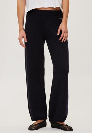 Personne portant un pantalon en maille noir ample avec des poignets élastiques, des ballerines en résille noire, un top court blanc et un bracelet doré, debout sur un fond clair.