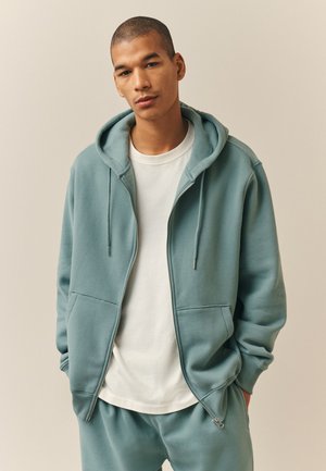 Mladý muž nosí tykysovú mikinu na zips a zodpovedajúce tepláky s bielym tričkom, stojí pred jednoduchým pozadím.