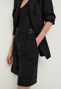 Zwarte, op maat gemaakte blazer en bijpassende shorts van gestructureerde stof, met plooien, een riemlus met een zilveren gesp en opgerolde zoom.