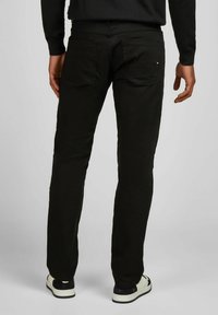 Homme portant un jean noir et des baskets noir et blanc, vu de dos contre un fond clair uni.