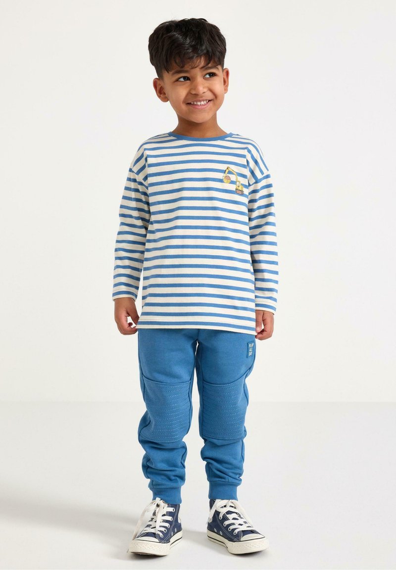 Koszula w paski z długim rękawem w kolorze kremowym i jasnoniebieskim, z małą grafiką. Niebieskie spodnie dresowe z teksturowanymi akcentami i ściągaczami przy kostkach. Białe trampki.