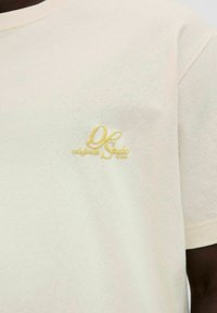 Svetlo bež bombažna majica s teksturirano površino, okroglim izrezom in majhnim rumenim vezenim logotipom na prsih.