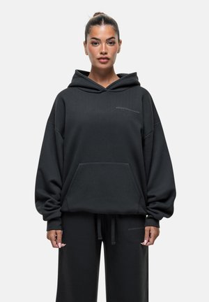 Zwarte oversized hoodie van zacht materiaal, met een kangoeroezak, trekkoord en subtiele reliëfdruk van het logo op de borst.