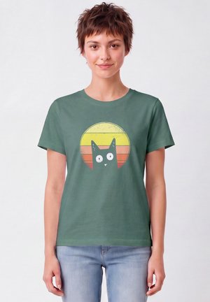 Jeune personne aux cheveux courts portant un t-shirt vert avec un chat noir dessin animé et un motif rétro de coucher de soleil jaune-orange, jean bleu clair.