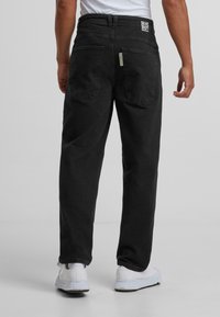 Pantalon en denim noir avec une coupe droite, dotée de deux poches arrière, d'un patch logo sur la ceinture et de détails de décoloration légère.