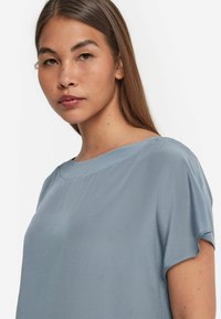 Ljusblå, kortärmad blus i mjukt och slätt material med rund halsringning och subtil glans; har en avslappnad passform.