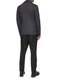 Dunkelblazer aus strukturiertem Stoff mit einem strukturierten Kragen und taillierter Silhouette. Kombiniert mit schwarzen Hosen und schwarzen Schuhen, von hinten betrachtet.