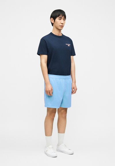 Νεαρός άνδρας που στέκεται φορώντας ανοιχτό μπλε αθλητικό σορτς, μπλε σκούρο μπλουζάκι με γραφικά, λευκές κάλτσες και λευκά αθλητικά παπούτσια, πάνω σε απλό φόντο.