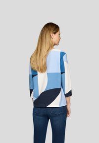 Blusenshirt mit blau-weißem Muster und 3/4-Ärmeln, das abstrakte Formen und eine strukturierte Oberfläche aufweist, kombiniert mit dunkelblauen Jeans.