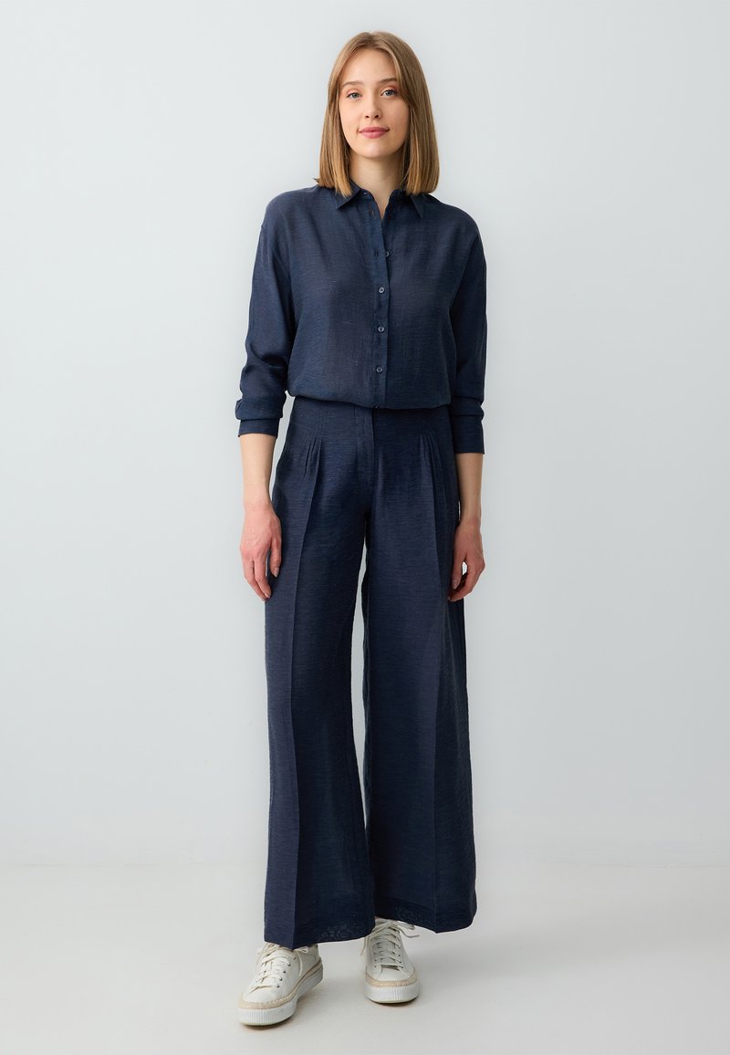 Marineblauw overhemd met lange mouwen, gecombineerd met wijde broekspijpen. Beide gemaakt van een gestructureerde stof, aangevuld met witte sneakers.