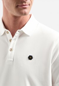 Witte polo met een textuurstof, voorzien van een zwart hexagonaal embleem met gele accenten en drie knopen bij de kraag.