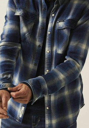 Person iført en blå og hvid ternet skjorte med knapper, justerer venstre manchet med begge hænder.