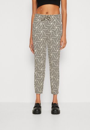 Pantalons blancs courts avec un motif géométrique noir, présentant une texture lisse, une taille mi-haute et des éléments de design angulaires. Des chaussures noires complètent le look.