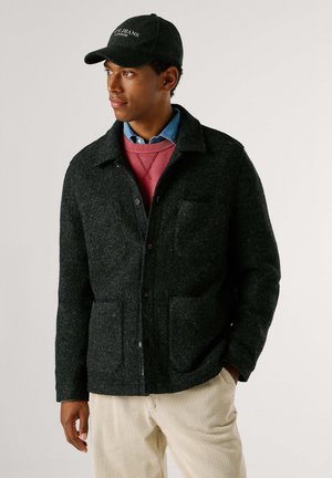 Hombre con gorra negra, chaqueta gris oscuro abotonada, sudadera roja, camisa azul y pantalones de pana beige, mirando hacia la izquierda sobre un fondo liso.