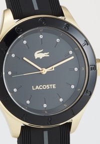 Montre Lacoste avec bracelet en caoutchouc noir, boîtier en acier inoxydable doré, cadran noir, index des heures en cristal et lettres gravées sur la lunette.
