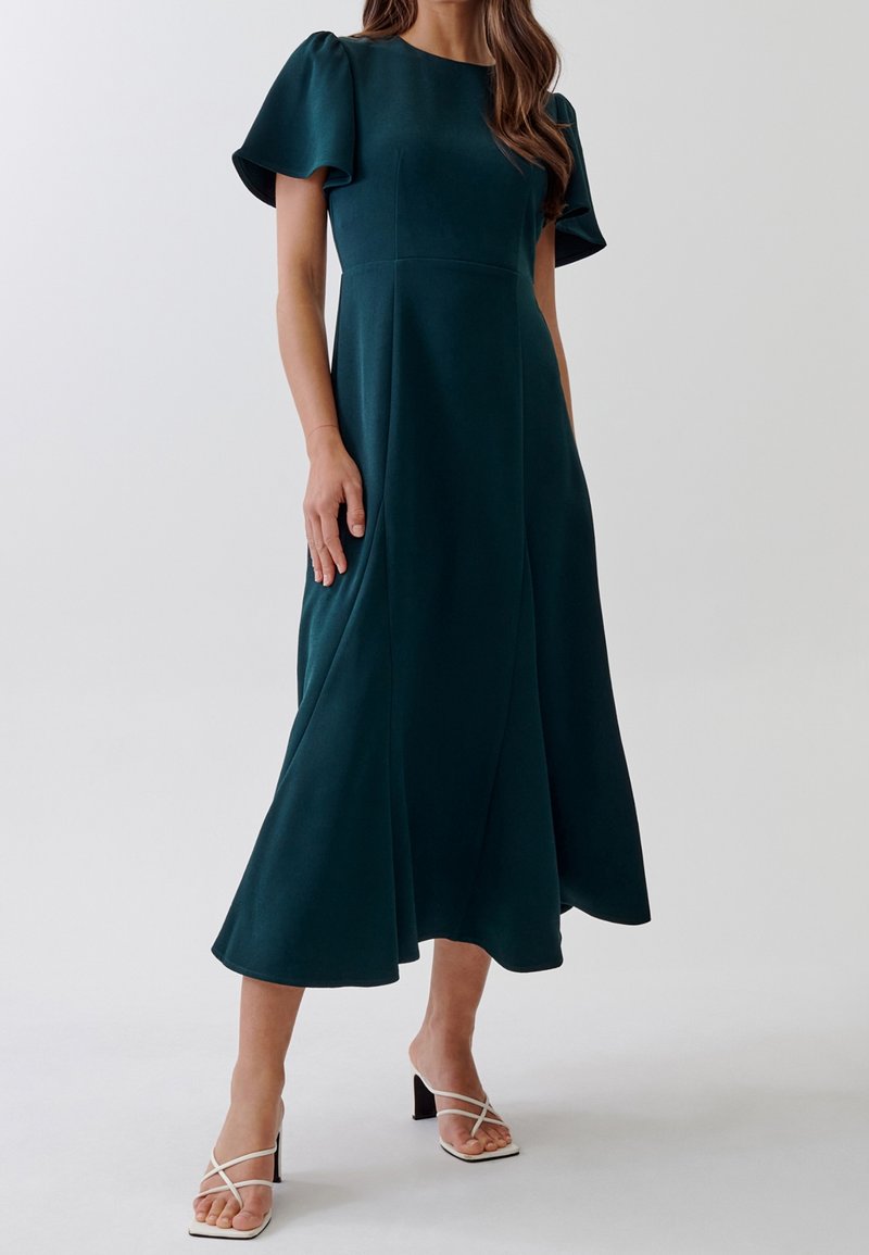 Femme portant une robe midi bleu sarcelle foncé à manches courtes volantées et des sandales blanches à talons et brides, debout devant un fond uni.