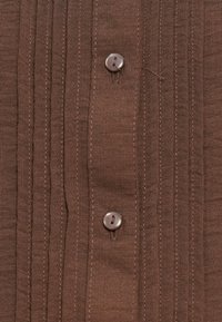 Camisa de algodão castanha com riscas verticais, dois botões e uma textura suave. O tecido apresenta detalhes de costura sutis.