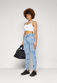 Reggiseno sportivo bianco, leggings azzurri tie-dye e scarpe da ginnastica nere. La modella tiene una borsa da palestra nera. Design semplice e tessuto traspirante.