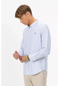 Giovane che sistema il polsino di una camicia a righe bianche e azzurre, indossando pantaloni beige e braccialetti colorati, su sfondo semplice.