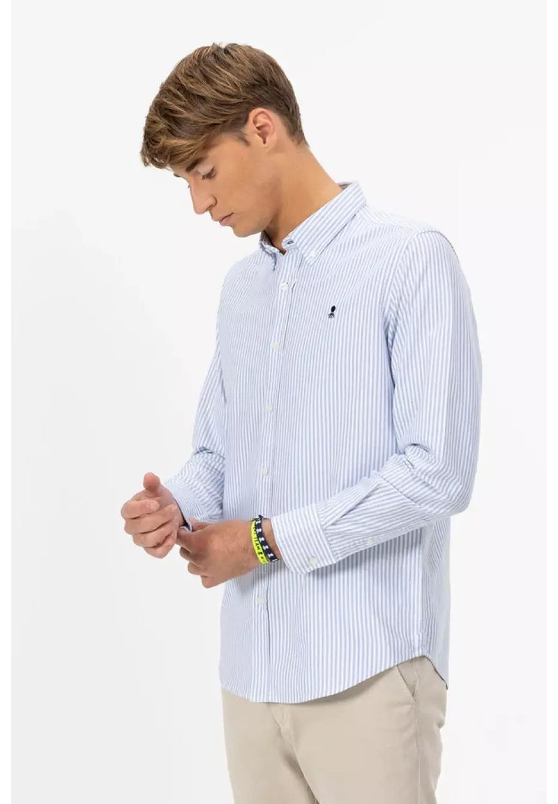 Giovane che sistema il polsino di una camicia a righe bianche e azzurre, indossando pantaloni beige e braccialetti colorati, su sfondo semplice.