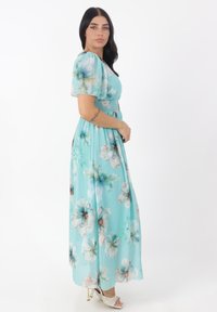 Ljusblå blommig maxiklänning med puffärmar, smockad midja och flödande tyg. Har stora pastellblommor och en mjuk textur.