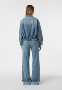 Kobieta w jasnoniebieskiej dżinsowej kurtce z tyłu i szerokich dżinsach z mankietami, stojąca na tle białym.