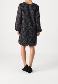 Black lace dress featuring floral pattern, long puffed sleeves, and a scalloped hem. Worn with black block-heeled shoes.