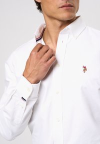 Valkoinen nappipaita, jossa on jämäkkä kaulus ja punainen, valkoinen ja sininen raita sisäpuolella. Rintaa koristaa kirjailtu logo. Sileä kangas.